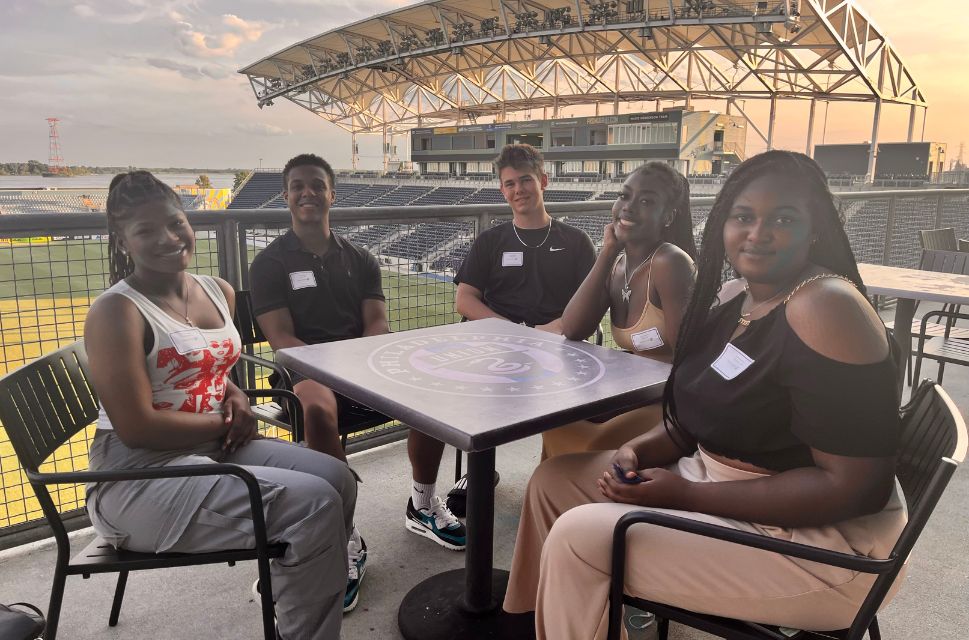 Five students seated at a table with the Subaru Park stadium as background