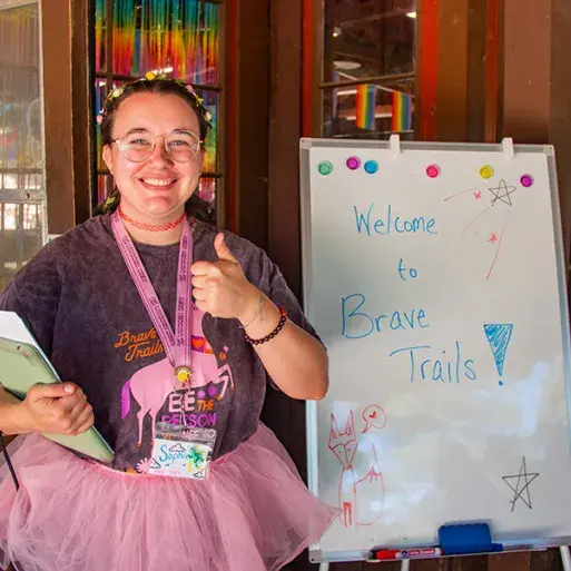 Sophia smiles at the camera giving a thumbs up. They wear a dark shirt and a pink tutu. Next to them is a white board with "Welcome to Brave Trails" written on it 