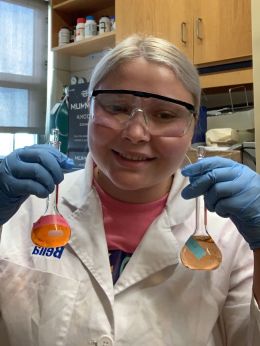 Student in white lab coat and goggles holds up two flasks filled with yellow-orange liquid