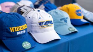 Four Widener University hats in different styles are lined up on blue tablecloth.