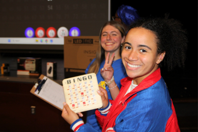 A student in the foreground holding a BINGO card and a student in background smiling