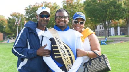 A mother and father stand on each side oft their son, smiling. He is wearing a band uniform with a Homecoming sash draped across him. They are standing outside on Widener's campus.