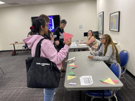 Three students stand at two tables engaging in an activity, as two other people sit behind the tables.