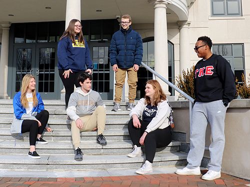 Six students stand or sit on the steps of Founders Hall 