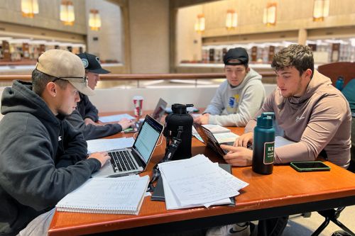 Four students studying at a table in the library with books, papers and laptops open