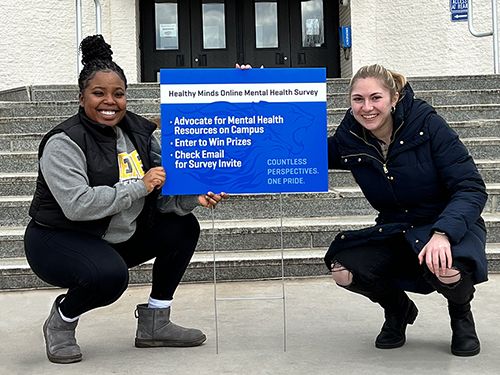 Two students kneel next to a blue lawn sign advertising a mental health survey